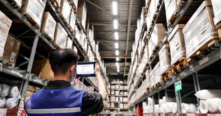 Warehouse worker using a rugged tablet for stock verification and inventory management in a high-rack warehouse.